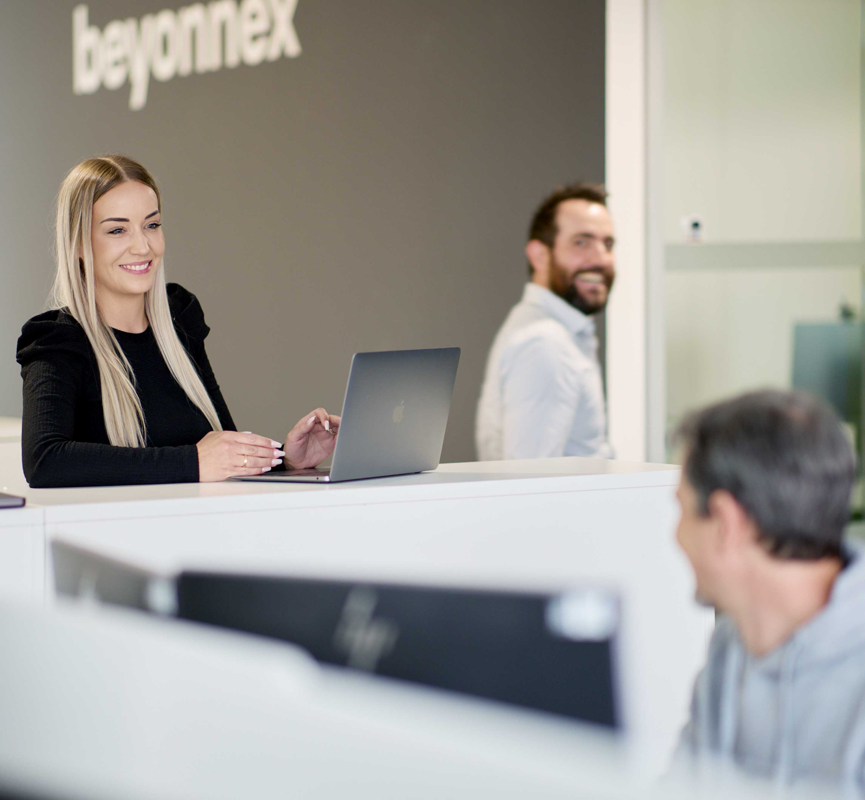 A woman with long blonde hair is smiling and working on a laptop at a standing desk in a modern office. Two men are interacting in the background, and the word 